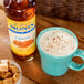 A blue mug with white liquid next to a brown bottle of Monin Sugar Free Caramel Flavoring Syrup.