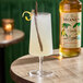 A glass of a light-colored beverage garnished with a vanilla bean and lemon twist, with a bottle of Monin Organic Vanilla Flavoring Syrup in the background.