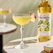 A bottle of Monin Premium Pear Flavoring Syrup next to a yellow cocktail garnished with a dried fruit slice.