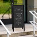 A black Aarco A-Frame chalkboard sign on the sidewalk with vegetables and prices.