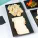 A black Cal-Mil faux slate rectangular platter with a raised rim holding cheese and crackers on a table.