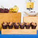 A Cal-Mil Madera pine rectangle plate riser holding two muffins in brown wrappers on a table with bowls of fruit.