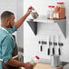 A man in a brown apron and blue shirt holding a bottle of brown powder over a stainless steel shelf.