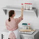 A woman in a white shirt standing on a Regency stainless steel wall shelf in a professional kitchen holding a container of spices.