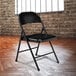 A National Public Seating black metal folding chair in front of a brick wall.