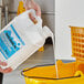 A hand pouring Sierra by Noble Chemical high performance floor finish from a white jug into a yellow bucket.