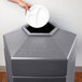 A hand putting a white plate into a gray hexagonal trash can.