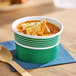 An 8 oz. green paper food cup filled with soup and bread pieces, placed on a blue napkin next to a wooden spoon.