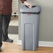 A man in a chef's uniform standing next to a Rubbermaid Untouchable gray square trash can.