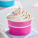 A pink paper frozen yogurt or food cup filled with swirled vanilla soft serve and colorful sprinkles.