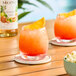 A glass of a tropical orange and red drink with ice and a pineapple wedge garnish, served on a wooden table.
