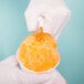 A gloved hand pours orange Carnival King snow cone syrup into a cup.