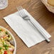 A white 2-ply dinner napkin with a fork placed on top, set on a wooden table.