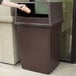 A person holding a brown Rubbermaid Glutton trash can outside.