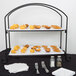 A black rectangular display stand with two tiers holding a tray of food with silver utensils.