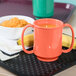 A green Rio orange two-handled mug filled with liquid on a table with a bowl of food.