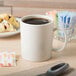 An ivory GET Tritan mug filled with brown liquid on a table with a spoon and a plate of pastries.