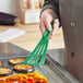 A hand holding a green Mercer Culinary Hell's Tools slotted spatula over a pan of vegetables.