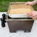 A person pouring food into a large metal pan in a Sterno Copper Vein fold away chafer on a table in an outdoor catering setup.