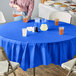 A woman sitting at an outdoor table with a Creative Converting cobalt blue tablecloth, and glasses of orange liquid.