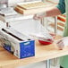 A woman in a green shirt and gloves using a slide cutter to wrap a bowl of red liquid with Choice Safecut premium foodservice film.