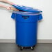 A woman using a blue Rubbermaid BRUTE trash can with a plastic lid to throw away a cardboard box.