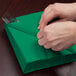 A person is wrapping a Creative Converting emerald green luncheon napkin around a fork and knife.