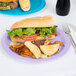 A sandwich with chips and a luscious lavender paper plate with a fork and knife on a table.