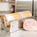 A roll of plastic wrap on a stainless steel Bulman countertop dispenser next to a roll of ham.