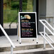 An Aarco aluminum A-Frame sidewalk sign with a black porcelain menu board that says "Taco" on the sidewalk outside of a restaurant.