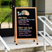 An Aarco solid oak A-frame sidewalk board with a black porcelain marker board on a sidewalk.