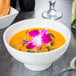 An American Metalcraft white porcelain footed bowl filled with soup and topped with a flower on a table with a white plate and spoon.