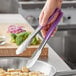 A person holding Vollrath stainless steel tongs with a purple Kool-Touch handle over a bowl of food.