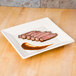 A Homer Laughlin Ivory square china plate with steak and sauce on it.