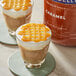 Two glasses of cappuccino with caramel topping next to a container of Ghirardelli Caramel Flavoring Sauce.