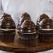 A chef holding a wood tray of chocolate cupcakes with Ghirardelli Dark Coating Wafers on a counter.