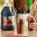 A gallon jug of Carnival King cola slush syrup concentrate next to two cups of prepared cola slushies with dome lids.