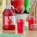 A gallon jug of Carnival King cherry slush syrup concentrate next to two cups of red cherry slushies with dome lids.