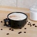 A black Tuxton jumbo cappuccino cup with white liquid and brown beans.
