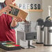 A woman pours coffee beans into a Bunn LPG coffee grinder on a counter in a coffee shop.