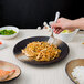 A person holding a fork over a 10 Strawberry Street Nagoya stoneware plate of noodles.