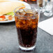 A GET SAN plastic stackable pint glass filled with soda and ice on a table with a sandwich.