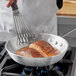 A hand using a spatula to cook meat in a Choice aluminum fry pan on a stove.