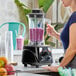 A commercial blender with a clear container filled with a purple smoothie, operated by a person in a kitchen setting.