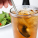 A hand holding a Libbey stainless steel iced tea spoon in a glass of ice tea.