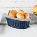 An oval Tablecraft rattan basket filled with rolls and fruit on a table.