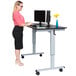 A woman standing at a black and gray Luxor stand up desk with a computer.