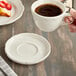 A person holding an Acopa ivory scalloped stoneware saucer with a cup of coffee on a wood table.