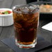 An Arcoroc stackable cooler glass filled with brown liquid and ice on a table with a bowl of food.