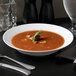 A white porcelain bowl of soup with a sprig of rosemary on a table.